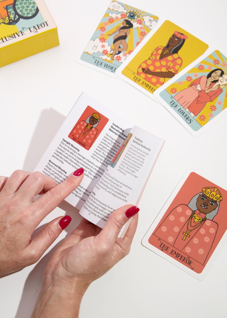 A pair of hands hold the karmic witch inclusive tarot deck guidebook open to read the meaning of the emperor card. Four major arcana cards can be seen in the background. 